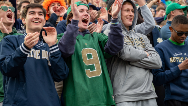 Three men wearing Notre Dame gear cheer and scream. A big crowd of Notre Dame fans can be seen in the back cheering on the football team.