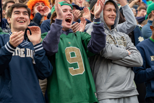 Three men wearing Notre Dame gear cheer and scream. A big crowd of Notre Dame fans can be seen in the back cheering on the football team.