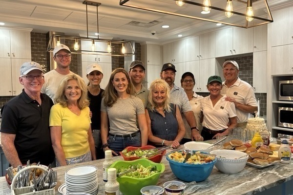 Ten individuals wearing Notre Dame-themed hats and attire gather behind a kitchen island laden with food. Bowls of salad, fruit salad, and hamburger buns suggest a casual gathering or cookout.