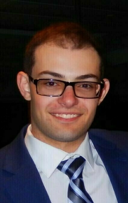 Headshot of a young man with short brown hair and black-rimmed glasses wearing a white shirt, blue and silver striped tie, and navy blue suit jacket, smiling at the camera.