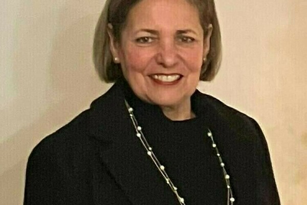 Woman with short graying hair, wearing a black turtleneck and jacket, smiles at the camera.  She wears a double-stranded pearl necklace.