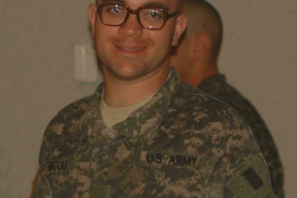 A man with a buzz cut, brown glasses, and a wide smile wears a U.S. Army camouflage uniform with "U.S. ARMY" and "NGOLL" name tapes. A blurry uniformed person stands behind him.