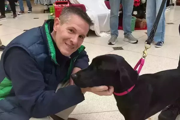 A smiling man in a blue and green vest crouches, gently petting a black dog wearing a bright pink collar and leash.