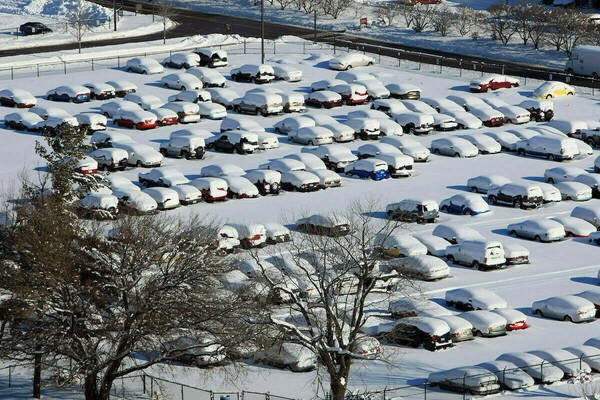 A vast Notre Dame parking lot blanketed in fresh white snow under a bright sun. Rows of cars are covered, revealing hints of red, blue, yellow, and dark colors. Bare trees dot the snowy landscape, highlighting a crisp winter day.