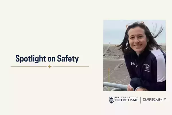 A smiling woman with dark hair and sunglasses on her head wears a navy and white Notre Dame athletic jacket, leaning on a railing with stadium seating behind her.