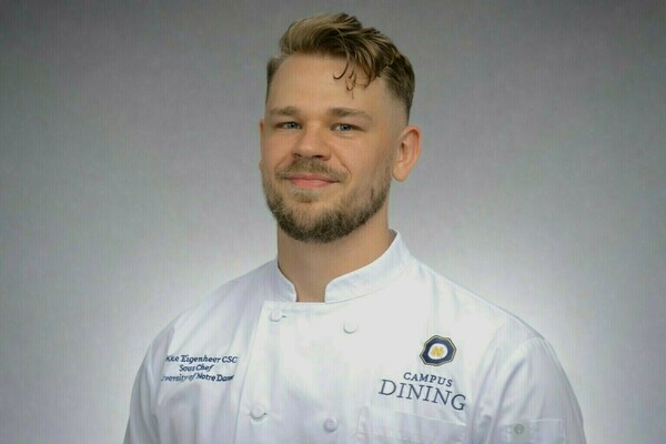 Jacob Eigenheer, Sous Chef at the University of Notre Dame, a young man with light brown hair and a beard, smiles while wearing a white chef's coat with the Campus Dining logo.