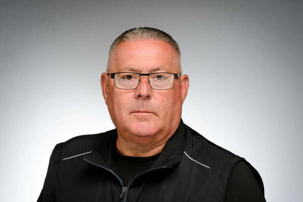 A man with white skin and short hair, with glasses, poses for a portrait. He is wearing a dark black zip up jacket.