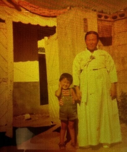 An adult in a light traditional robe stands with a young child in denim overalls, both smiling. The child holds hands to ears.