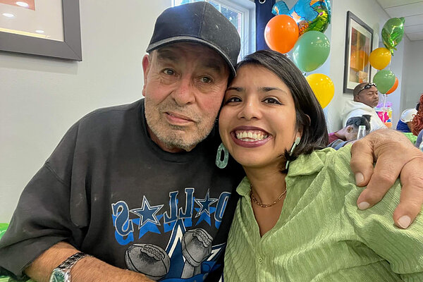 Elderly man in black baseball cap and woman in green shirt smile, embracing.