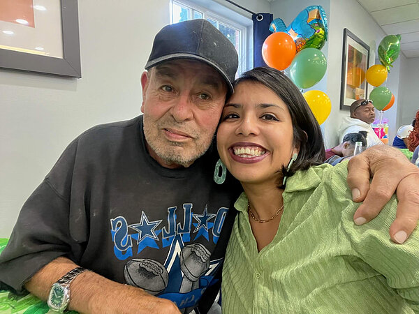 Elderly man in black baseball cap and woman in green shirt smile, embracing.